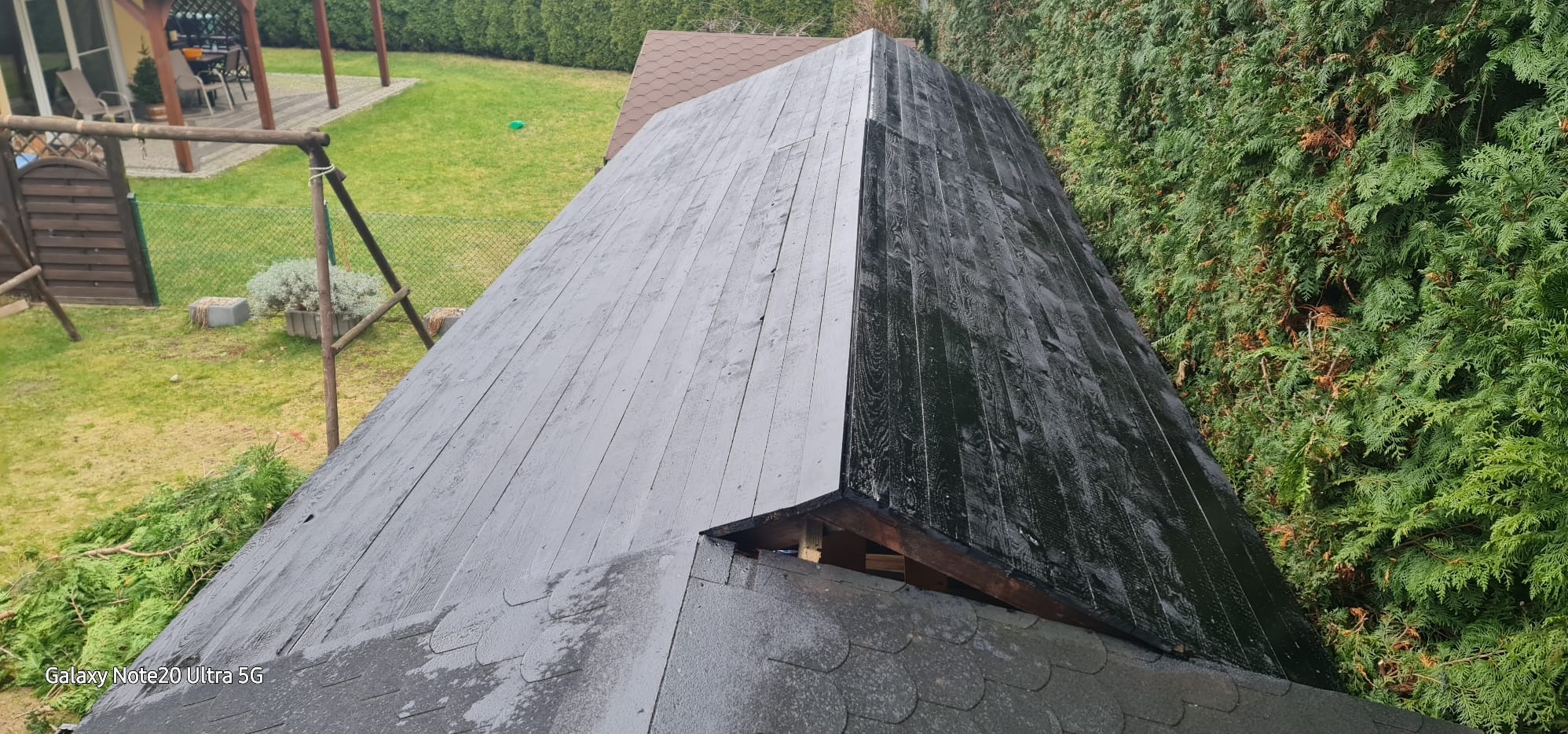 Fragment dachu po malowaniu: połowa w szarym kolorze, druga w czarnym. Widoczne deski i fragment ogrodu z zielonymi krzewami. Nietypowe ujęcie po renowacji.