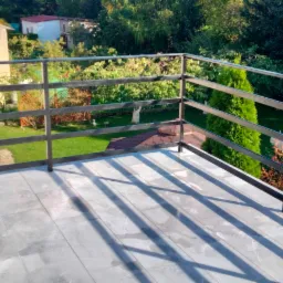 Nowoczesny balkon z szarymi płytkami i czarną, metalową balustradą, widok na ogród z zielonym trawnikiem i drzewami.