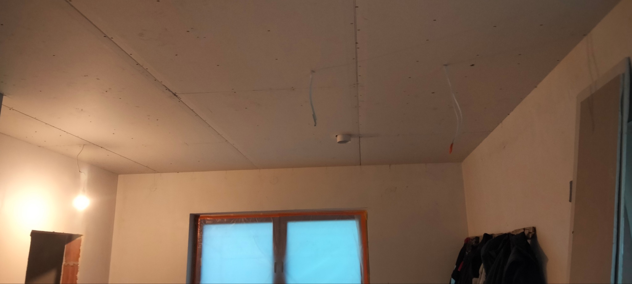 Sufit z płyt kartonowo-gipsowych, widoczne łączenia i wkręty, przewody wystające z sufitu, okno zabezpieczone folią malarską, ściany przygotowane do malowania.