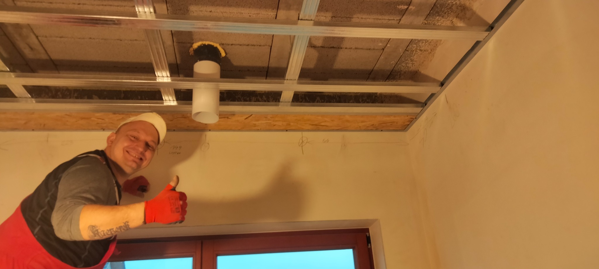 Uśmiechnięty monter w czerwonych rękawicach pokazuje kciuk w górę na tle metalowego rusztu sufitowego, częściowo zamontowanej płyty OSB i instalacji wentylacyjnej w trakcie prac wykończeniowych.