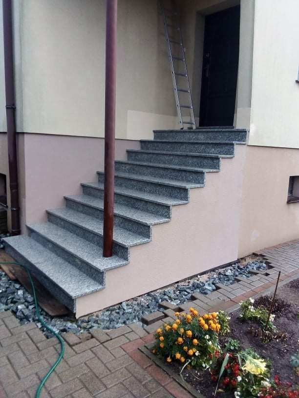 Zewnętrzne schody wejściowe wyłożone szarym kamieniem, z widoczną drabiną opartą o ścianę budynku i fragmentem ogrodu z kwiatami.