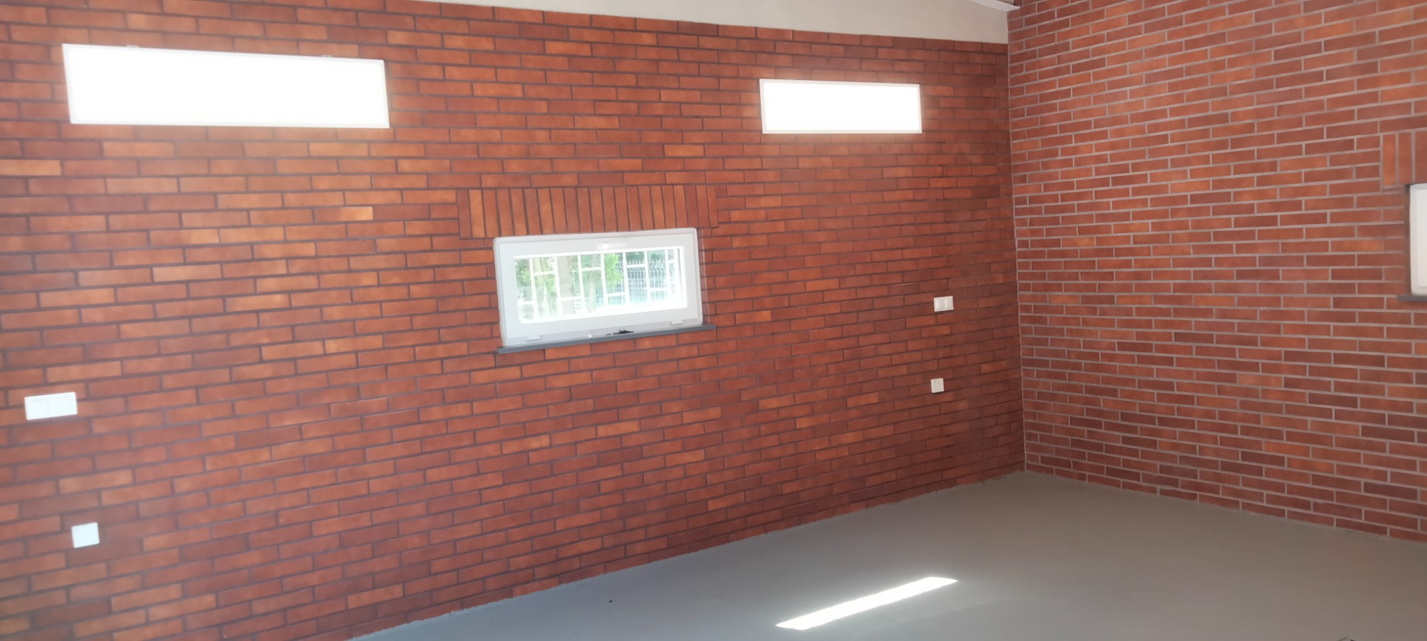 Wnętrze wykończone czerwoną cegłą klinkierową, widoczne okno z kratą, dwa prostokątne panele oświetleniowe i szara podłoga z delikatnym połyskiem.