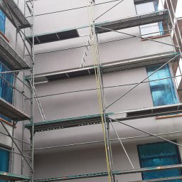 Uslugi - Budynek w trakcie prac elewacyjnych, widoczne rusztowania, okna zabezpieczone folią, świeżo nałożona warstwa tynku w odcieniach szarości.