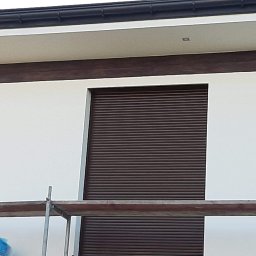 Uslugi - Fragment elewacji budynku z brązowymi roletami zewnętrznymi, widoczne rusztowanie i świeżo pomalowana biała ściana.