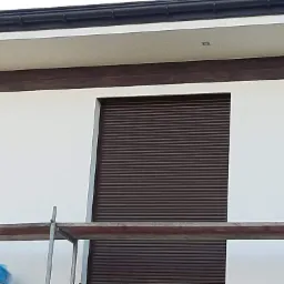 Fragment elewacji budynku z brązowymi roletami zewnętrznymi, widoczne rusztowanie i świeżo pomalowana biała ściana.