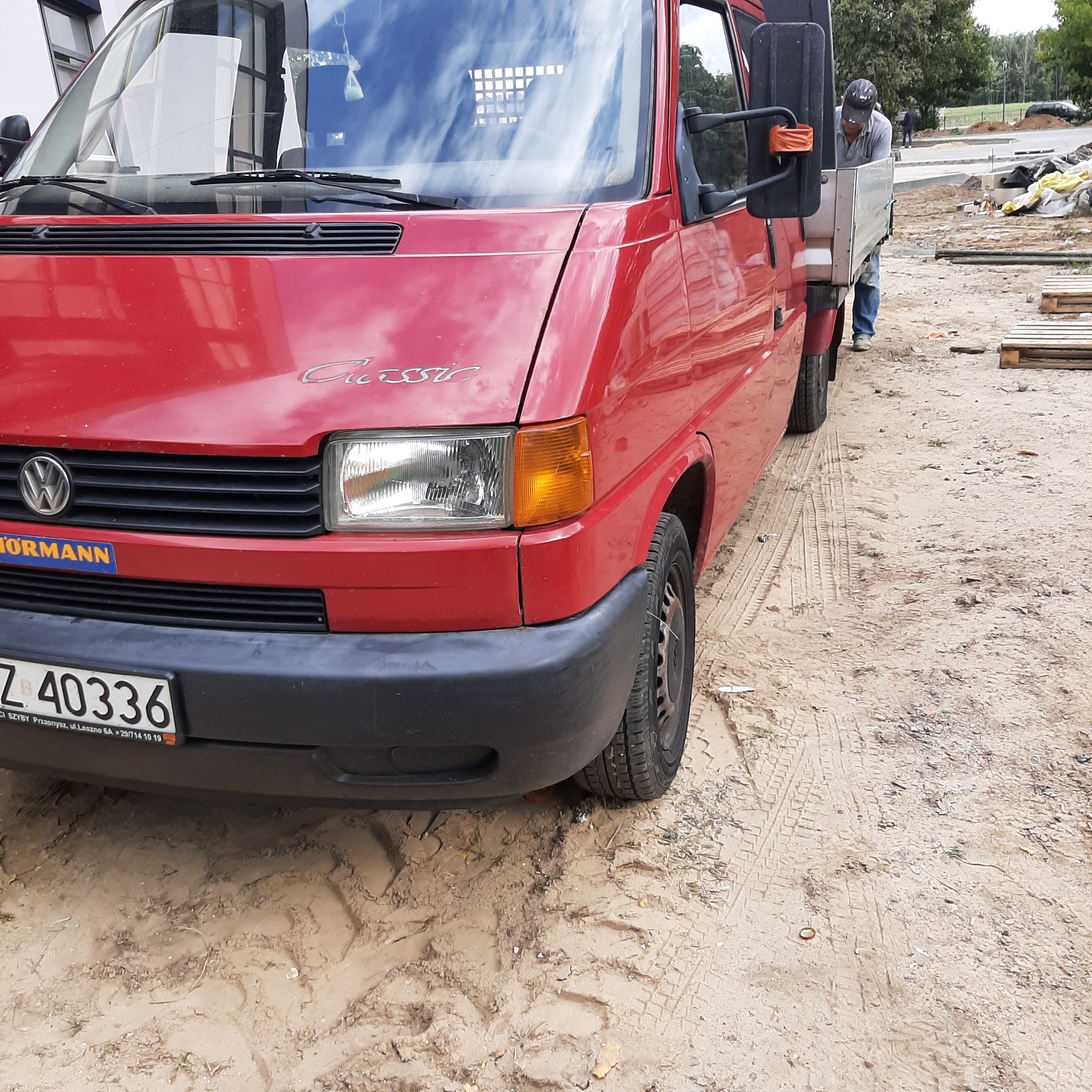 Czerwony samochód dostawczy Volkswagen Classic na piaszczystym terenie budowy, z pracownikiem w tle, widok z przodu na reflektor i tablicę rejestracyjną.
