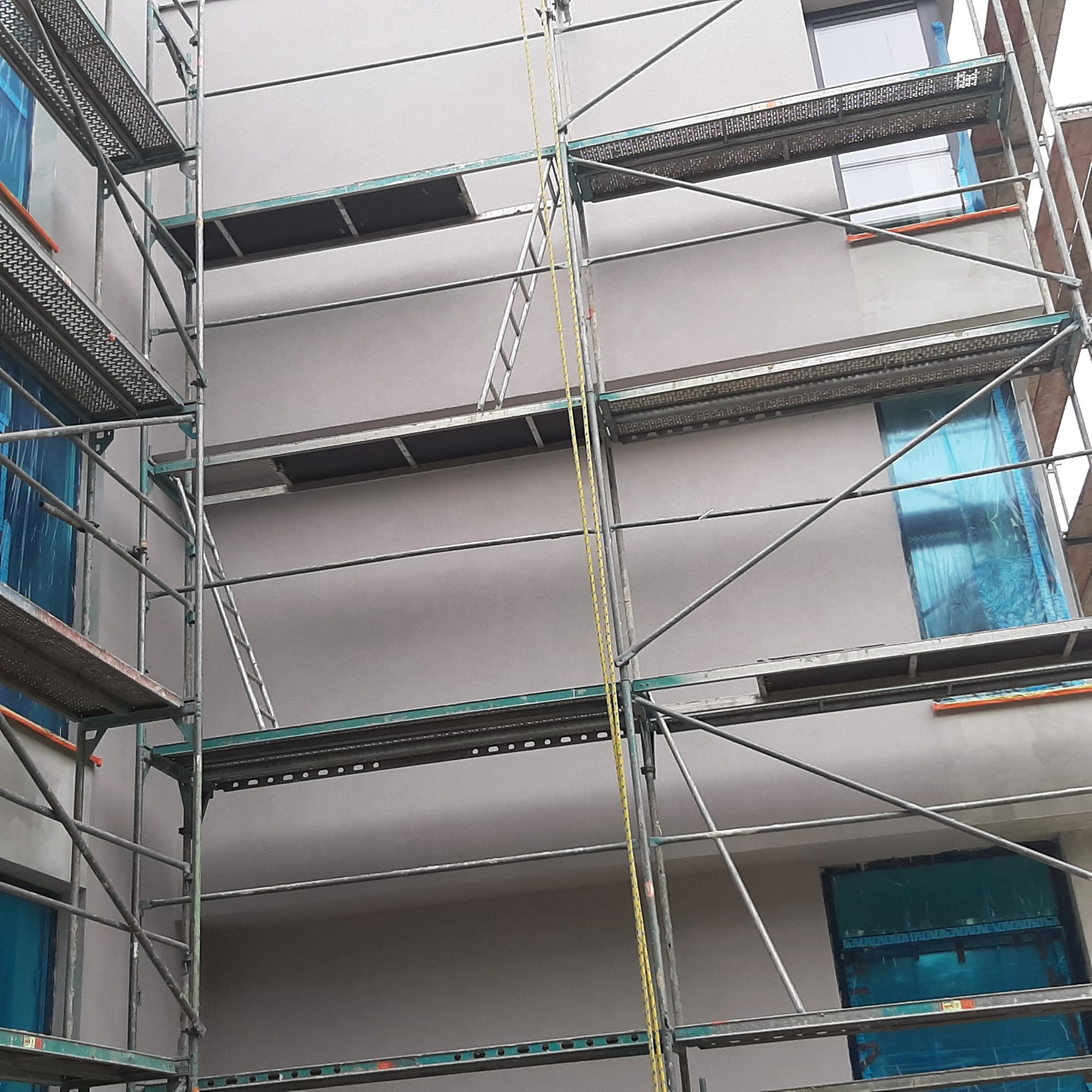 Budynek w trakcie prac elewacyjnych, widoczne rusztowania, okna zabezpieczone folią, świeżo nałożona warstwa tynku w odcieniach szarości.