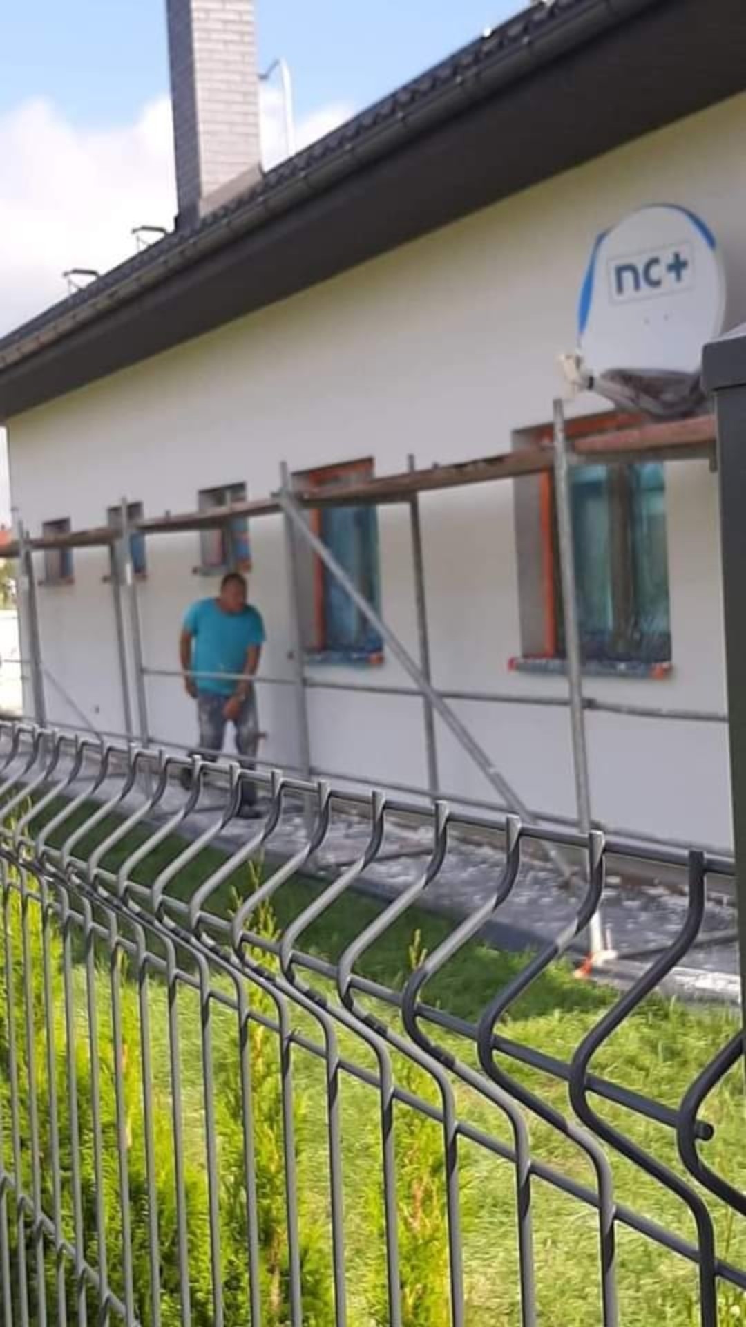 Pracownik w niebieskiej koszulce stoi na rusztowaniu przed ocieplonym domem z białą elewacją, widoczne zabezpieczone okna i antena satelitarna na ścianie, ujęcie zza metalowego ogrodzenia.