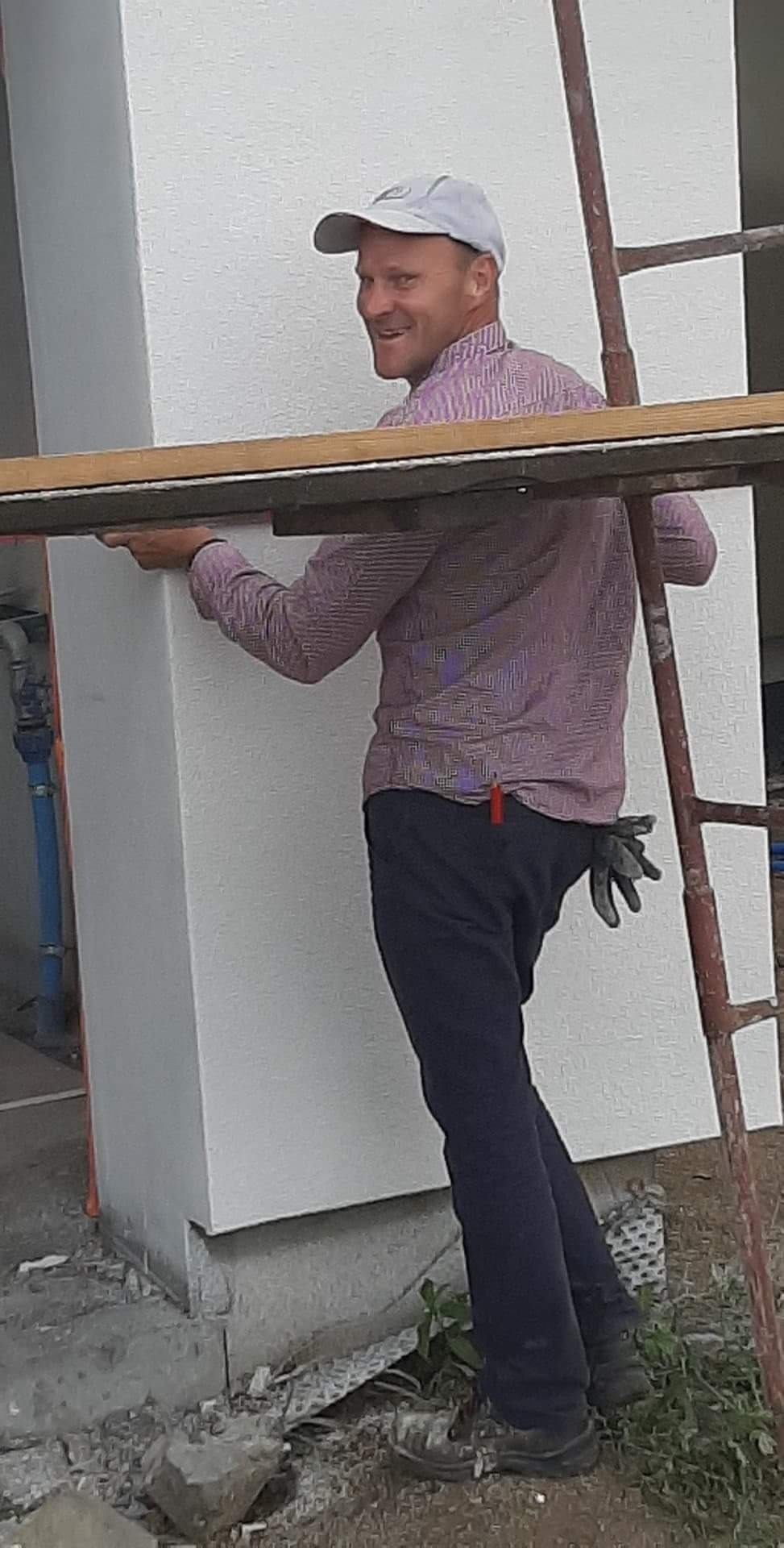 Uśmiechnięty mężczyzna w czapce i koszuli w paski, stojący na rusztowaniu przy ocieplonej ścianie budynku, z rękawicami w kieszeni spodni.