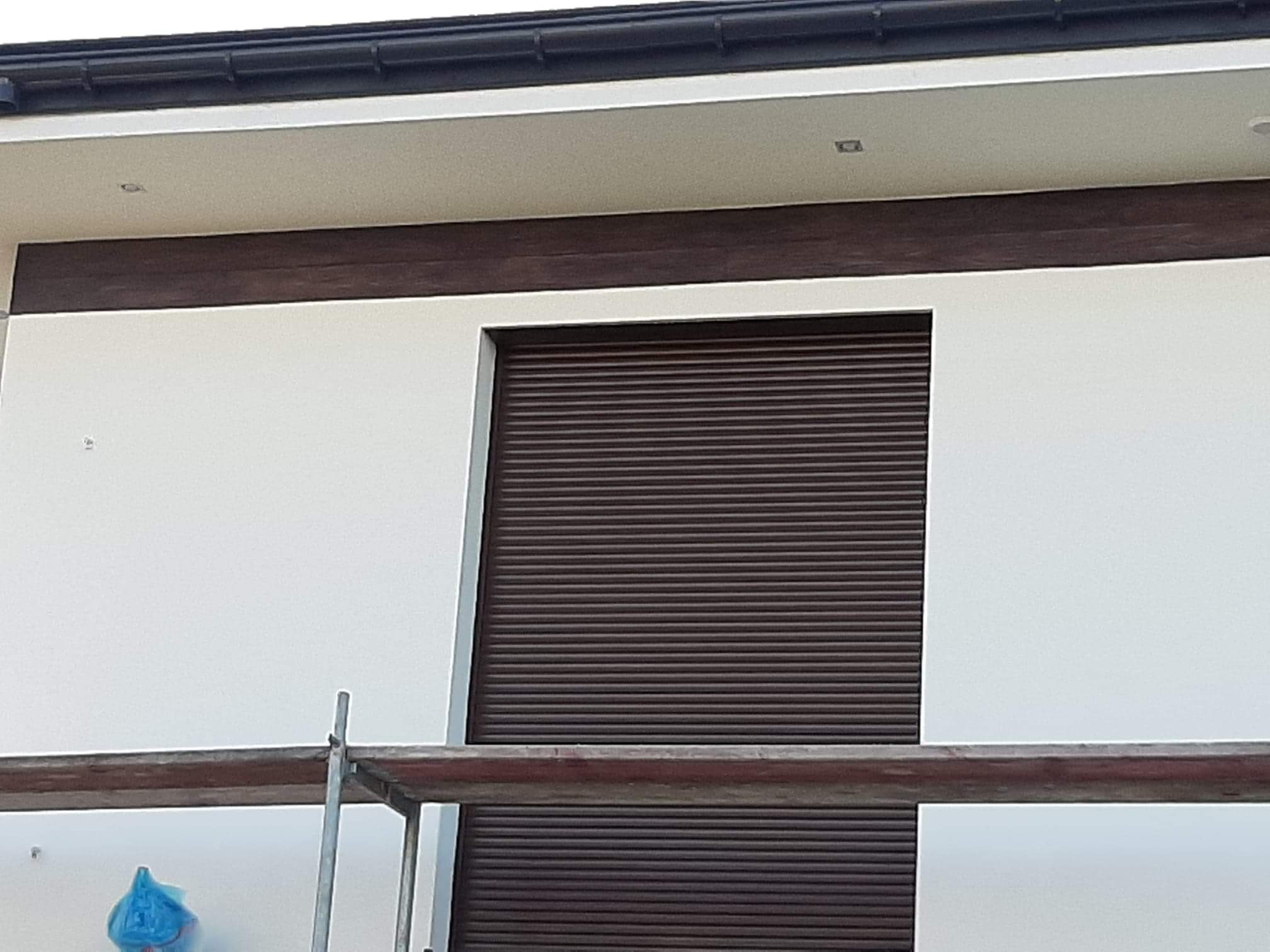 Fragment elewacji budynku z brązowymi roletami zewnętrznymi, widoczne rusztowanie i świeżo pomalowana biała ściana.