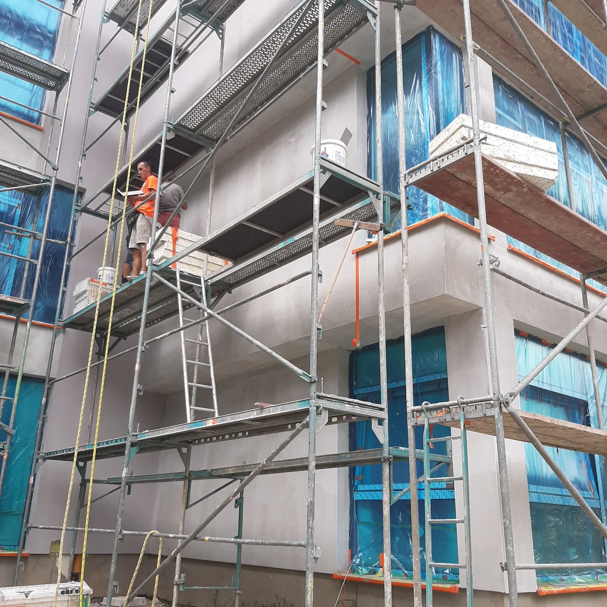 Pracownik na rusztowaniu sprawdza postęp prac na elewacji budynku, okna zabezpieczone folią, widoczne materiały izolacyjne i narzędzia.