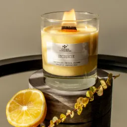 Płonąca sojowa świeca w szklanym pojemniku z drewnianym knotem, etykietą 'Szczęście' i plasterkiem cytryny, ustawiona na drewnianym podeście na lustrzanej powierzchni, dekorowana gałązką z drobnymi...