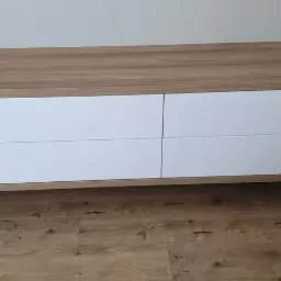 Wisząca komoda z czterema białymi szufladami i korpusem w kolorze jasnego drewna, zamontowana na ścianie nad podłogą z paneli w odcieniach brązu.