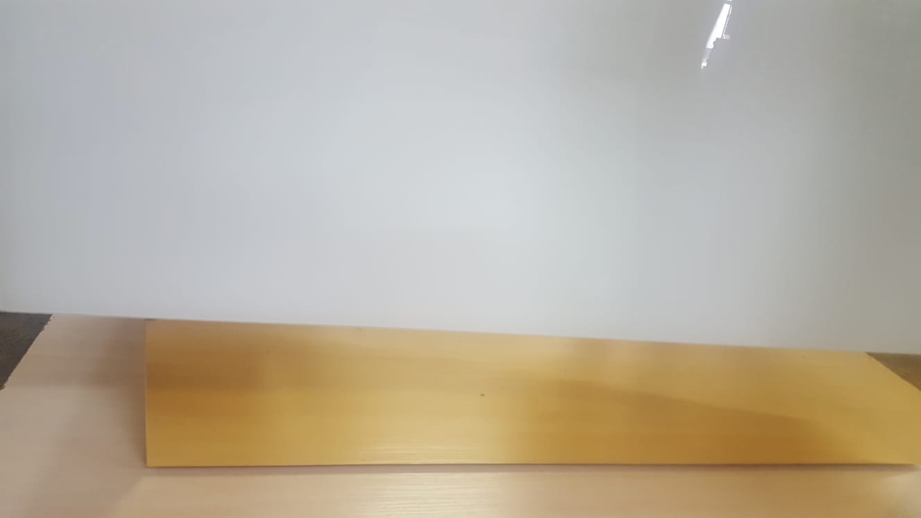 Jasny, drewniany blat o gładkiej powierzchni, leżący na podłożu, z widocznym fragmentem białej ściany w tle, oświetlony z góry.