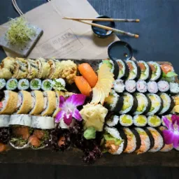 Bogato zdobiona, drewniana deska z różnorodnym sushi, w tym maki, nigiri i california rolls, udekorowana jadalnymi kwiatami orchidei i imbirem, z widoczną papierową torbą i pałeczkami w tle.