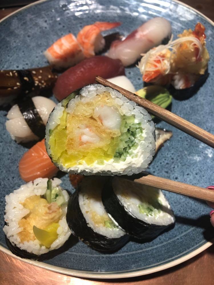 Różnorodny zestaw sushi na niebieskim talerzu, z bliska widoczne rolki z krewetką, ogórkiem i żółtą rzodkwią trzymane pałeczkami, obok nigiri z łososiem, węgorzem i tuńczykiem.