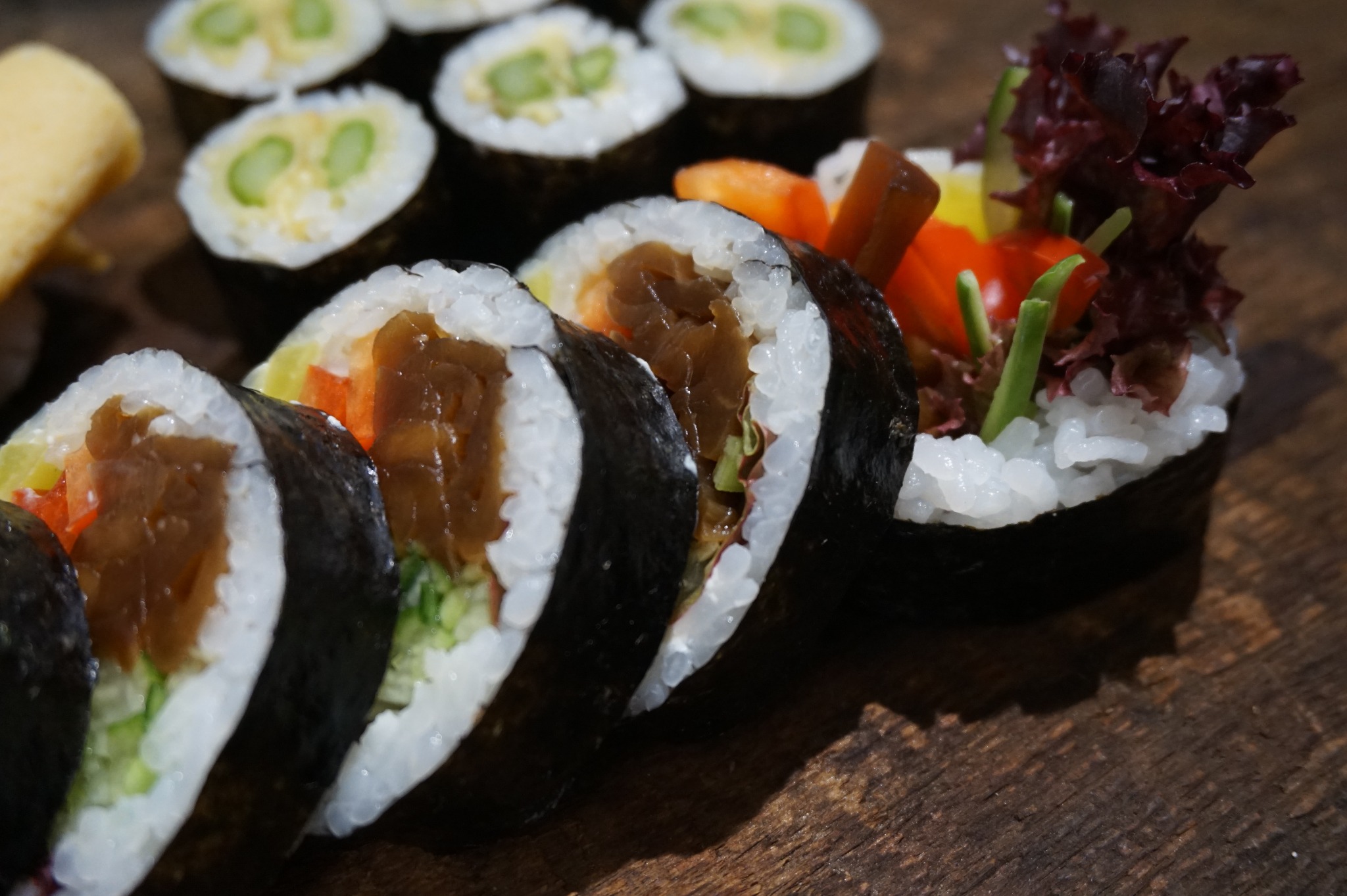 Ujęcie z bliska różnorodnych rolek sushi z wyraźnie widocznymi składnikami: brązowe grzyby, świeże warzywa i szparagi, ułożonych na drewnianej desce.