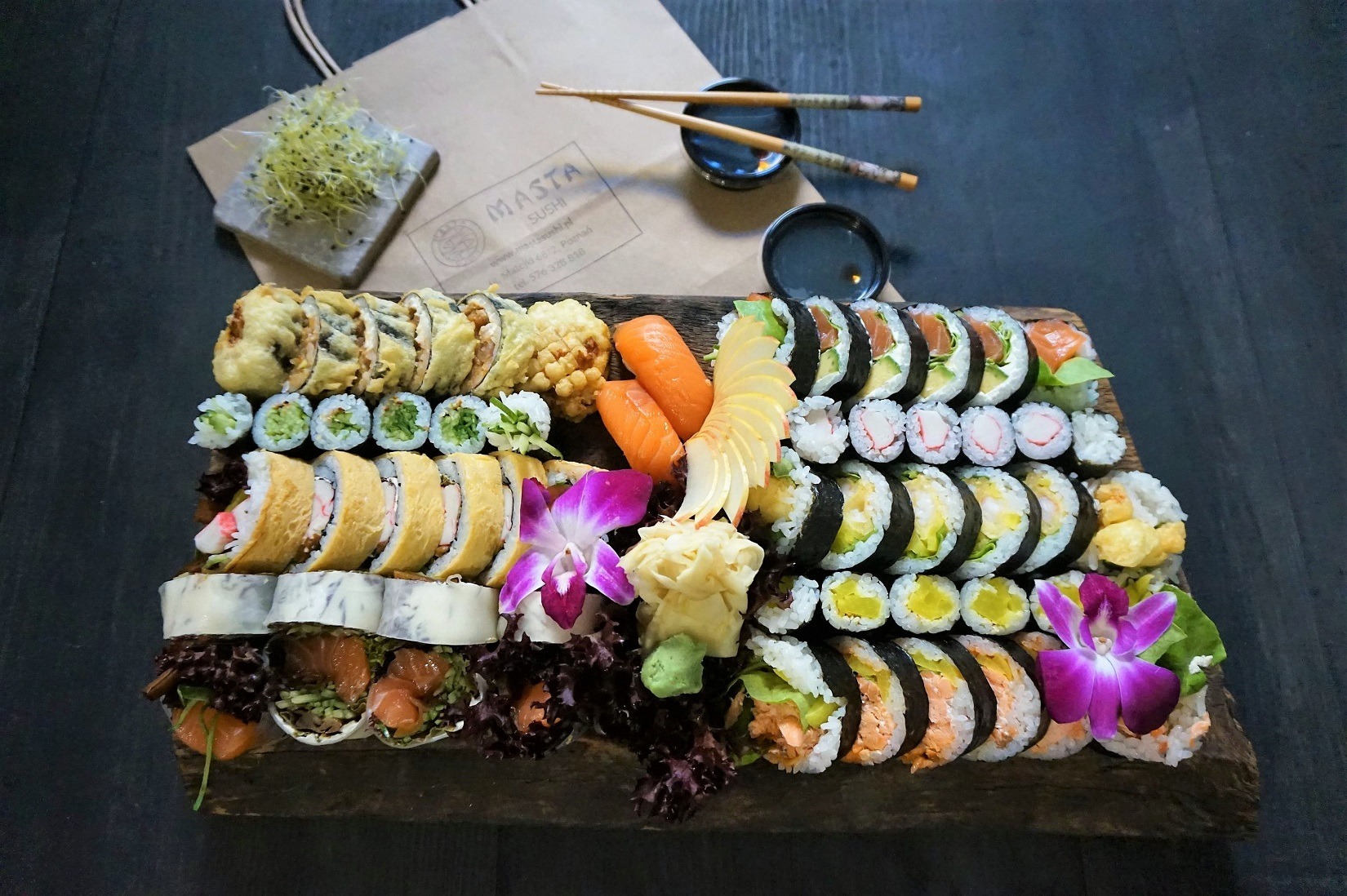 Bogato zdobiona, drewniana deska z różnorodnym sushi, w tym maki, nigiri i california rolls, udekorowana jadalnymi kwiatami orchidei i imbirem, z widoczną papierową torbą i pałeczkami w tle.