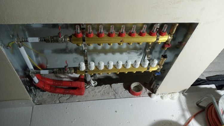 Rozdzielacz instalacji centralnego ogrzewania z rurami, zaworami i izolacją termiczną w szarej wnęce. Widoczne elementy montażowe i taśma izolacyjna.