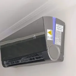 Czarny klimatyzator ścienny z wyświetlaczem temperatury, zamontowany w pomieszczeniu o białych ścianach i suficie. Widoczne naklejki informacyjne na obudowie.