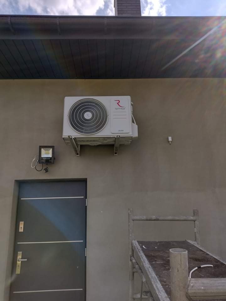 Biały klimatyzator z widocznym logo na szarej ścianie zewnętrznej domu, zamontowany nad drzwiami, obok lampy zewnętrznej, z widocznym fragmentem rusztowania.