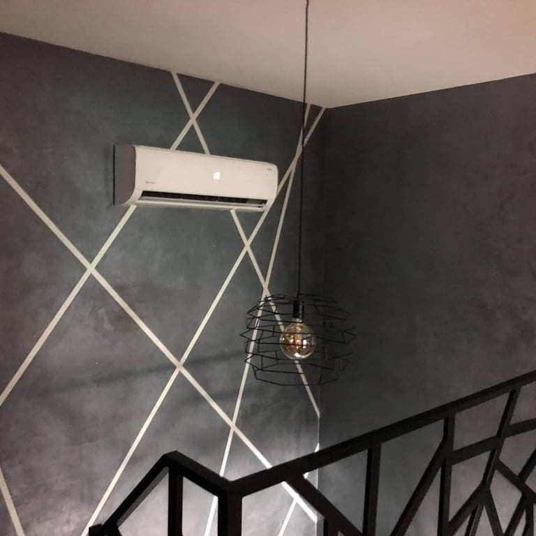 Nowoczesny klimatyzator ścienny zamontowany w pomieszczeniu o szarej ścianie z geometrycznym wzorem z białych linii, obok zwisa czarna lampa druciana z żarówką Edisona.