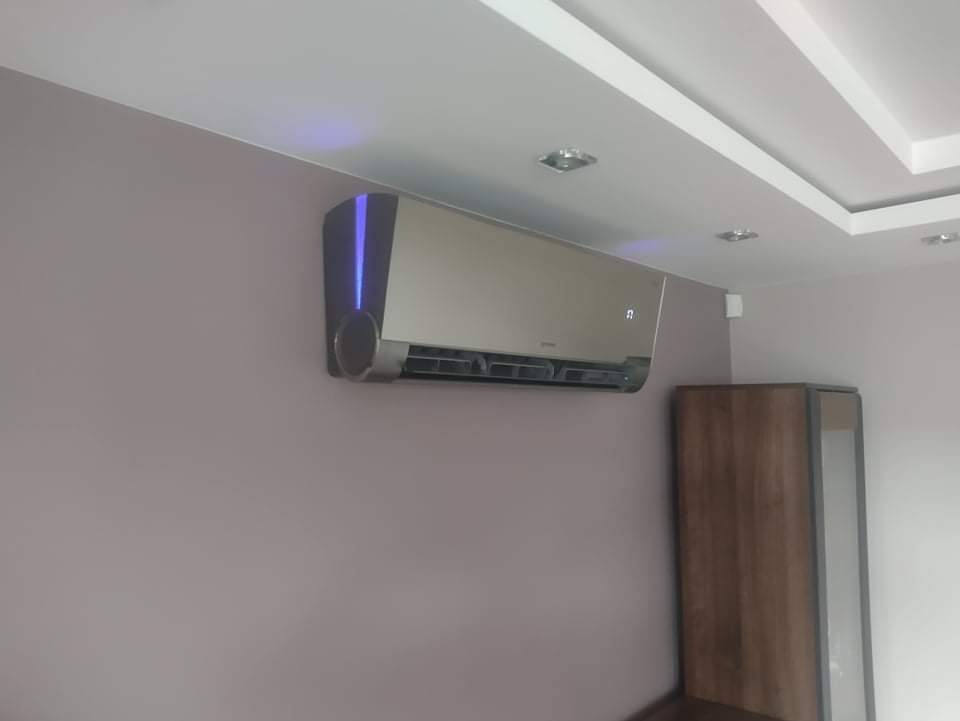 Nowoczesny klimatyzator ścienny w kolorze beżowym z niebieskim podświetleniem LED zamontowany w pomieszczeniu z szarą ścianą i białym sufitem.