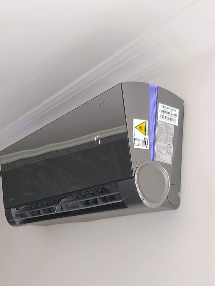 Czarny klimatyzator ścienny z wyświetlaczem temperatury, zamontowany w pomieszczeniu o białych ścianach i suficie. Widoczne naklejki informacyjne na obudowie.