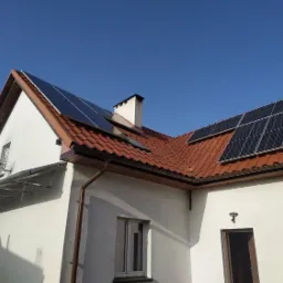 Biały dom z czerwoną dachówką i panelami słonecznymi na dachu w słoneczny dzień. Widoczne metalowe konstrukcje na ścianie domu.