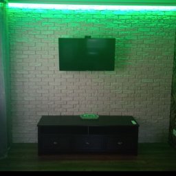 Alfa stroy bud - Ściana z cegieł dekoracyjnych podświetlona zielonym LED, z zawieszonym telewizorem i czarną szafką RTV.