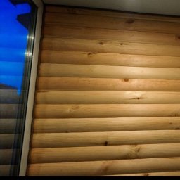 Alfa stroy bud - Ściana wykończona jasnym drewnem imitującym bale, z widocznym oknem i fragmentem niebieskiego nieba za nim, oświetlona górnym światłem.