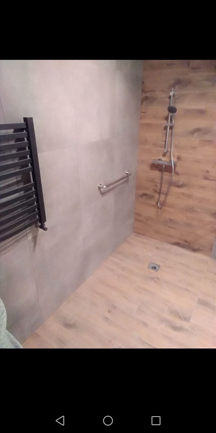 Nowoczesna kabina prysznicowa walk-in z drewnopodobnymi płytkami na ścianie i podłodze, chromowaną armaturą i grzejnikiem drabinkowym w łazience wykończonej szarymi płytkami.