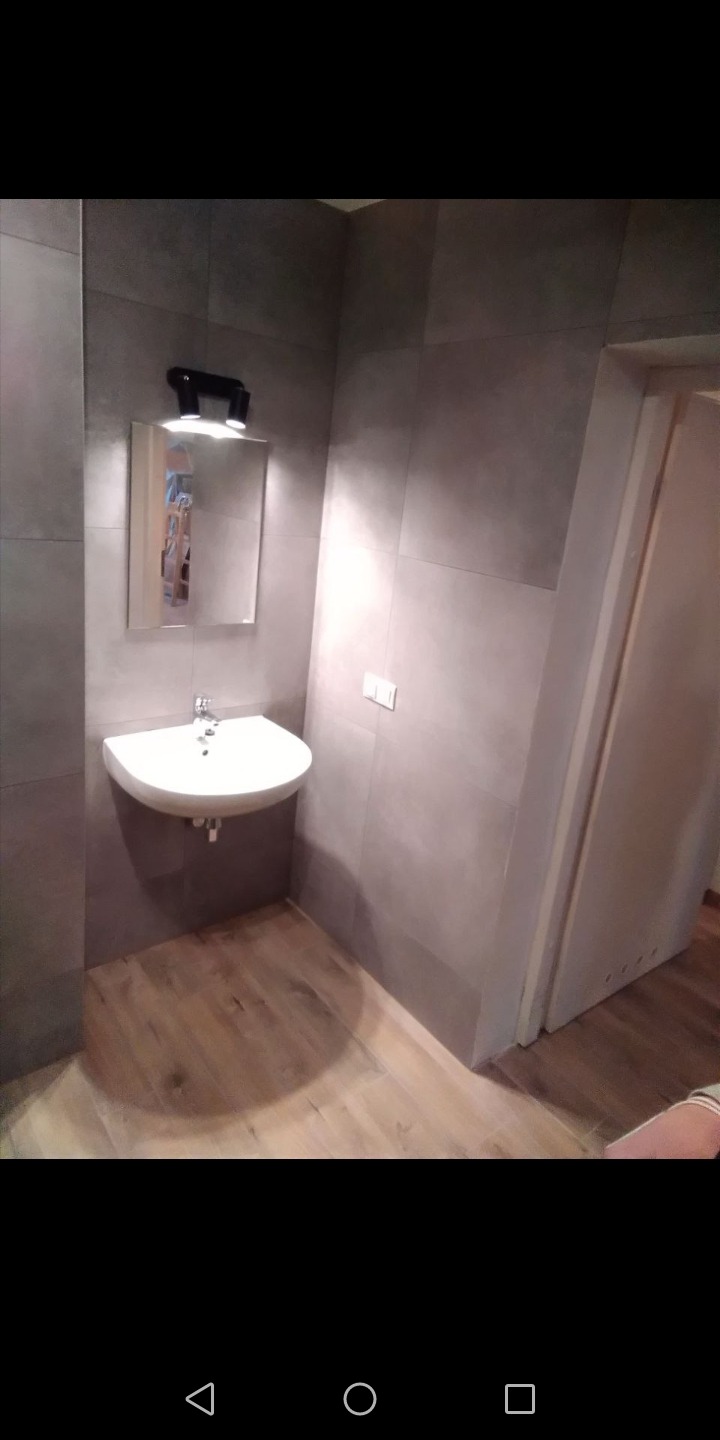 Umywalka narożna z lustrem podświetlanym dwoma reflektorami w łazience wykończonej szarymi płytkami i drewnopodobną podłogą.