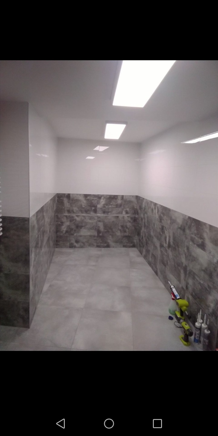 Wykończone wnętrze łazienki z szarymi płytkami imitującymi kamień na podłodze i dolnej części ścian, białe płytki na górze, widoczne narzędzia i tuby silikonu w rogu.