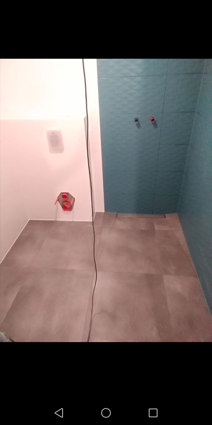 Wykończona łazienka z szarymi płytkami podłogowymi i turkusowymi płytkami ściennymi, widoczne przyłącza wodne i odpływ liniowy w podłodze, kabel zwisający z góry.