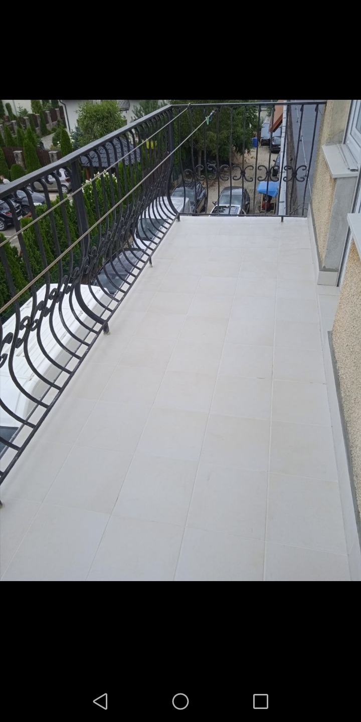 Białe płytki gresowe ułożone na podłodze balkonu z czarną, ozdobną balustradą; widok z góry na okoliczne drzewa i zaparkowane samochody.