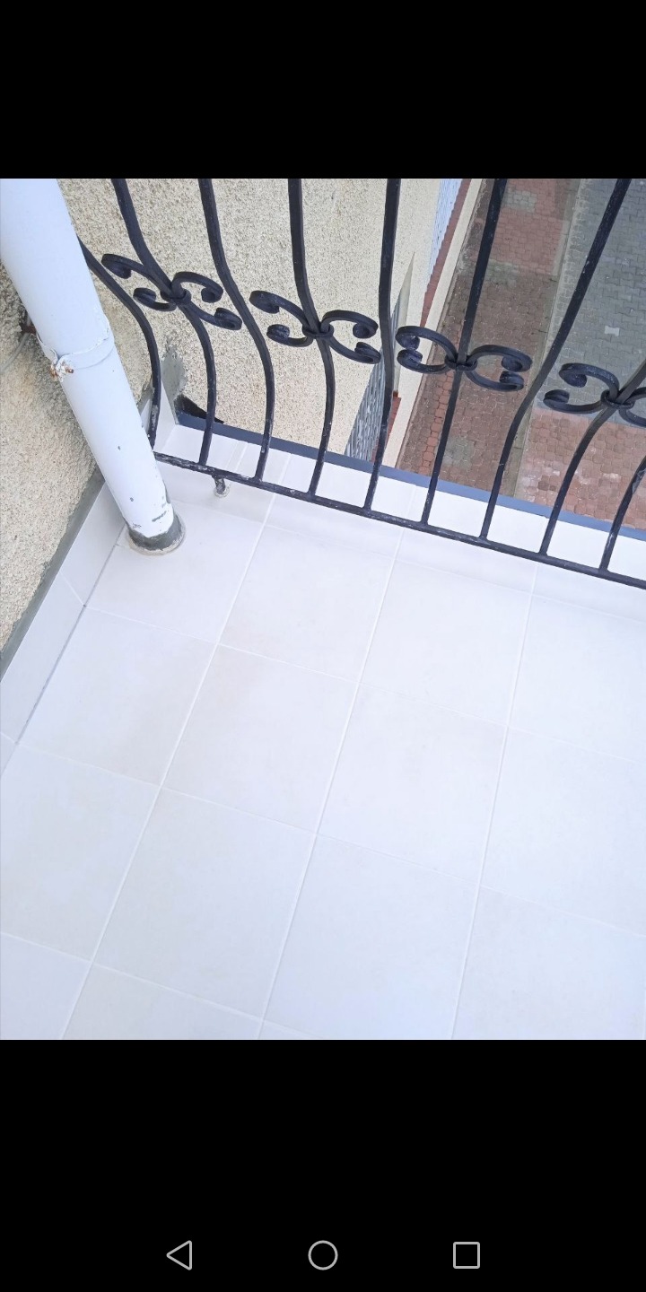 Białe płytki na balkonie z czarną, ozdobną balustradą i rurą spustową, widok z góry na fragment elewacji budynku z cegły.