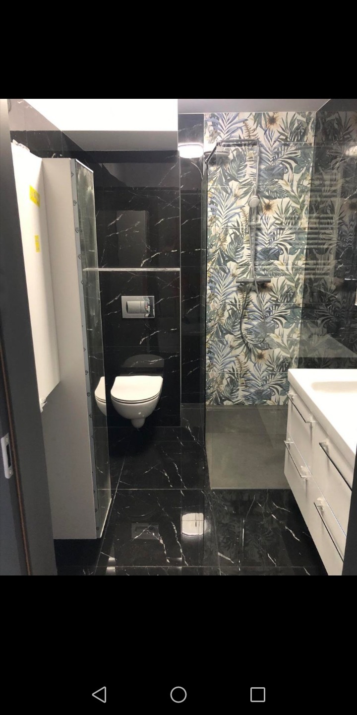 Nowoczesna łazienka z czarnymi marmurowymi płytkami, wiszącą toaletą i kabiną prysznicową z motywem liści, częściowo widoczną szafką umywalkową z białymi frontami.