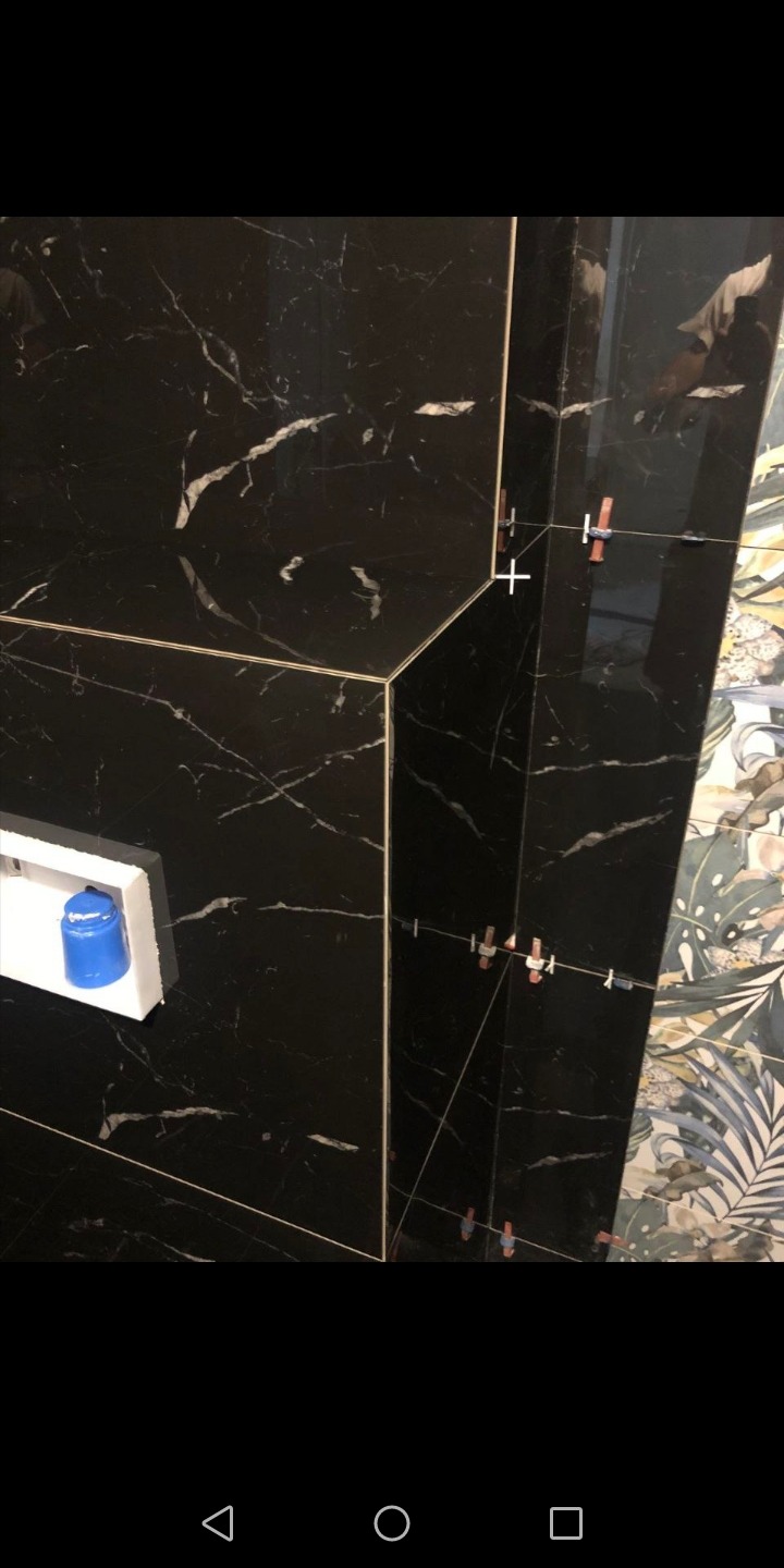 Czarne płytki imitujące marmur z białymi żyłami, ułożone na ścianie w rogu pomieszczenia, z widocznymi krzyżykami dystansowymi i niebieskim dozownikiem mydła w tle.