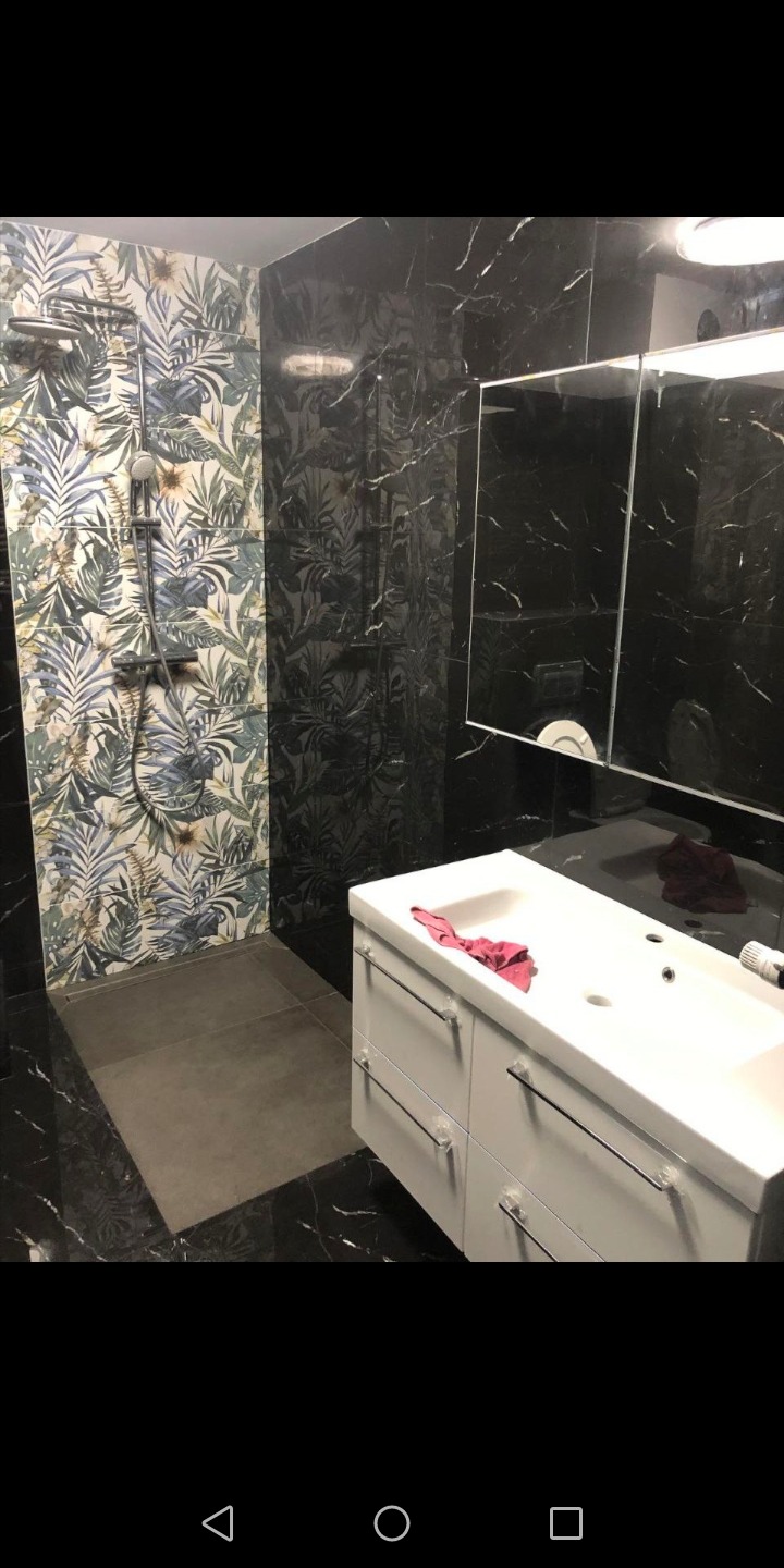 Wykończona łazienka z kabiną prysznicową w egzotyczne wzory i czarnymi marmurkowymi płytkami, biała szafka pod umywalką z pozostawioną czerwoną szmatką.