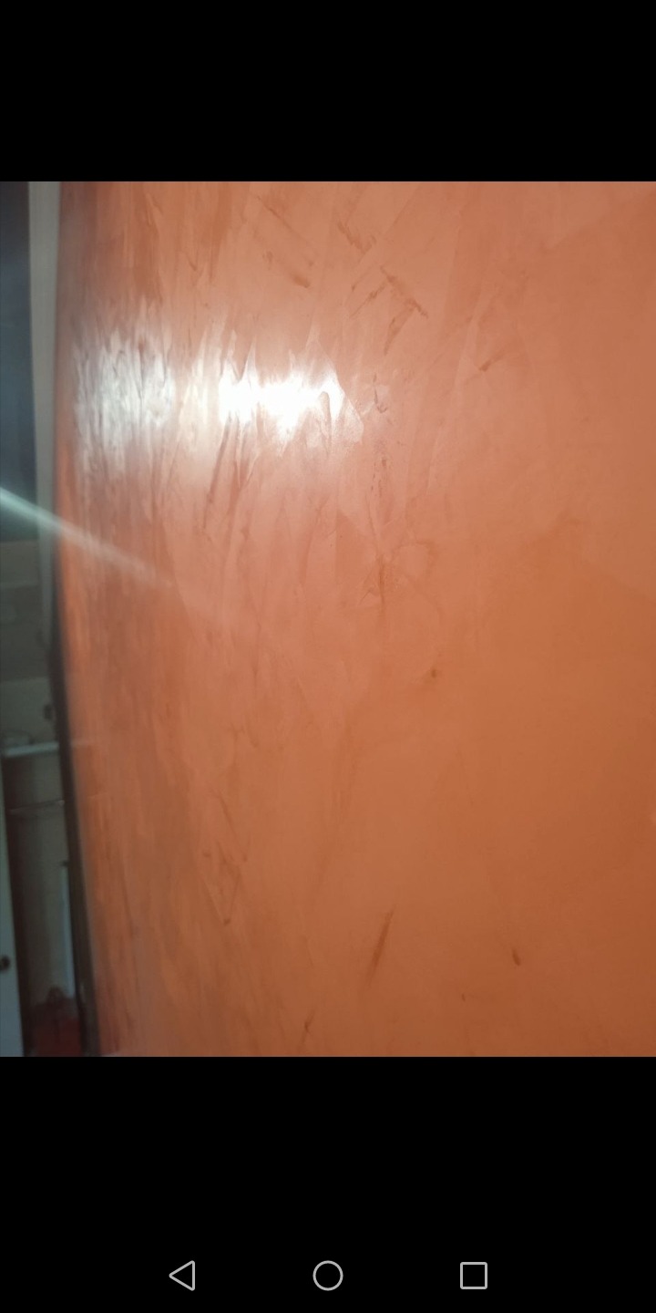 Ściana pokryta dekoracyjną, pomarańczową gładzią wenecką z widocznymi smugami i refleksami światła, oświetlona z boku, z fragmentem białej futryny drzwi po lewej stronie.