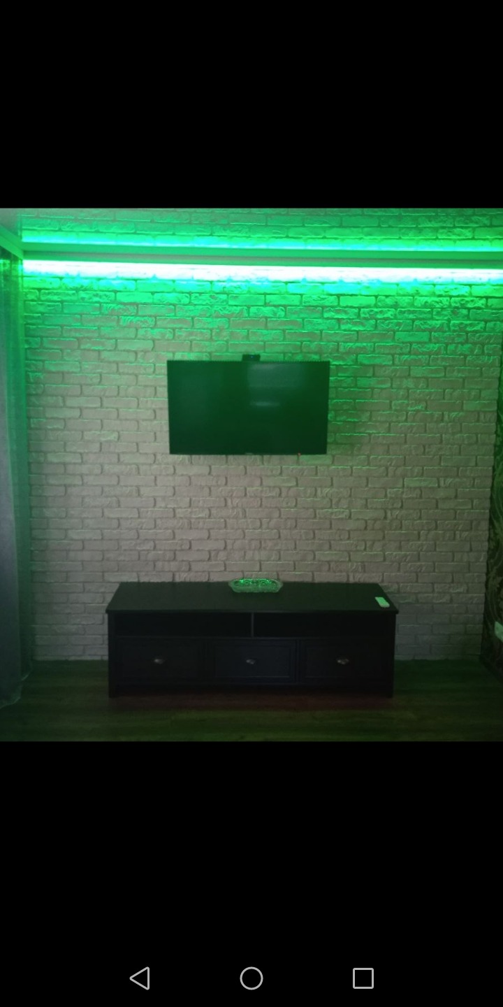 Ściana z cegieł dekoracyjnych podświetlona zielonym LED, z zawieszonym telewizorem i czarną szafką RTV.