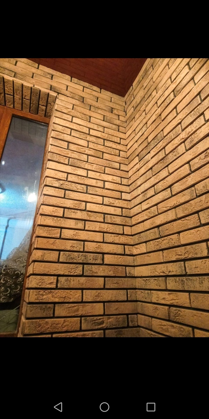 Wewnętrzny narożnik pomieszczenia wykończony dekoracyjną cegłą w odcieniach beżu i brązu, z widocznym fragmentem okna po lewej stronie i brązowym sufitem w górnym rogu.