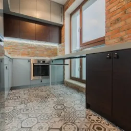 Nowoczesna kuchnia z szarymi szafkami i blatem, brązowymi frontami szafek wiszących, ceglaną ścianą i wzorzystą podłogą z płytek heksagonalnych.