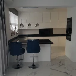 Nowoczesna kuchnia z białymi meblami na wymiar, czarnymi blatami, marmurową podłogą i dwoma granatowymi krzesłami barowymi z ćwiekami.