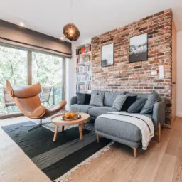 Salon z ceglaną ścianą, szarą sofą z szezlongiem, skórzanym fotelem BoConcept, okrągłym stolikiem z owocami i wzorzystym dywanem na jasnej drewnianej podłodze.