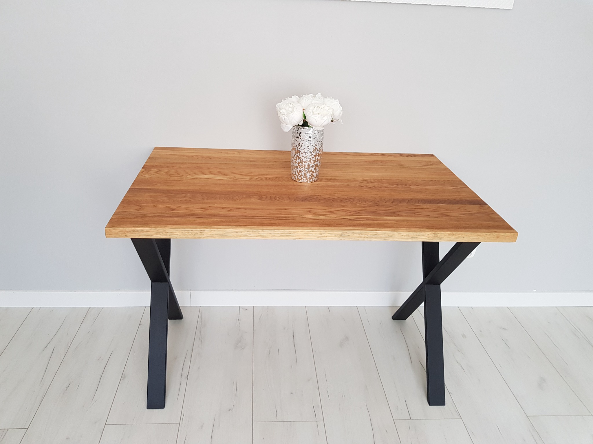 Stół z blatem z jasnego drewna o wyraźnym usłojeniu, z czarnymi, metalowymi nogami w kształcie litery X, na blacie srebrny wazon z bukietem białych kwiatów, na jasnej podłodze.