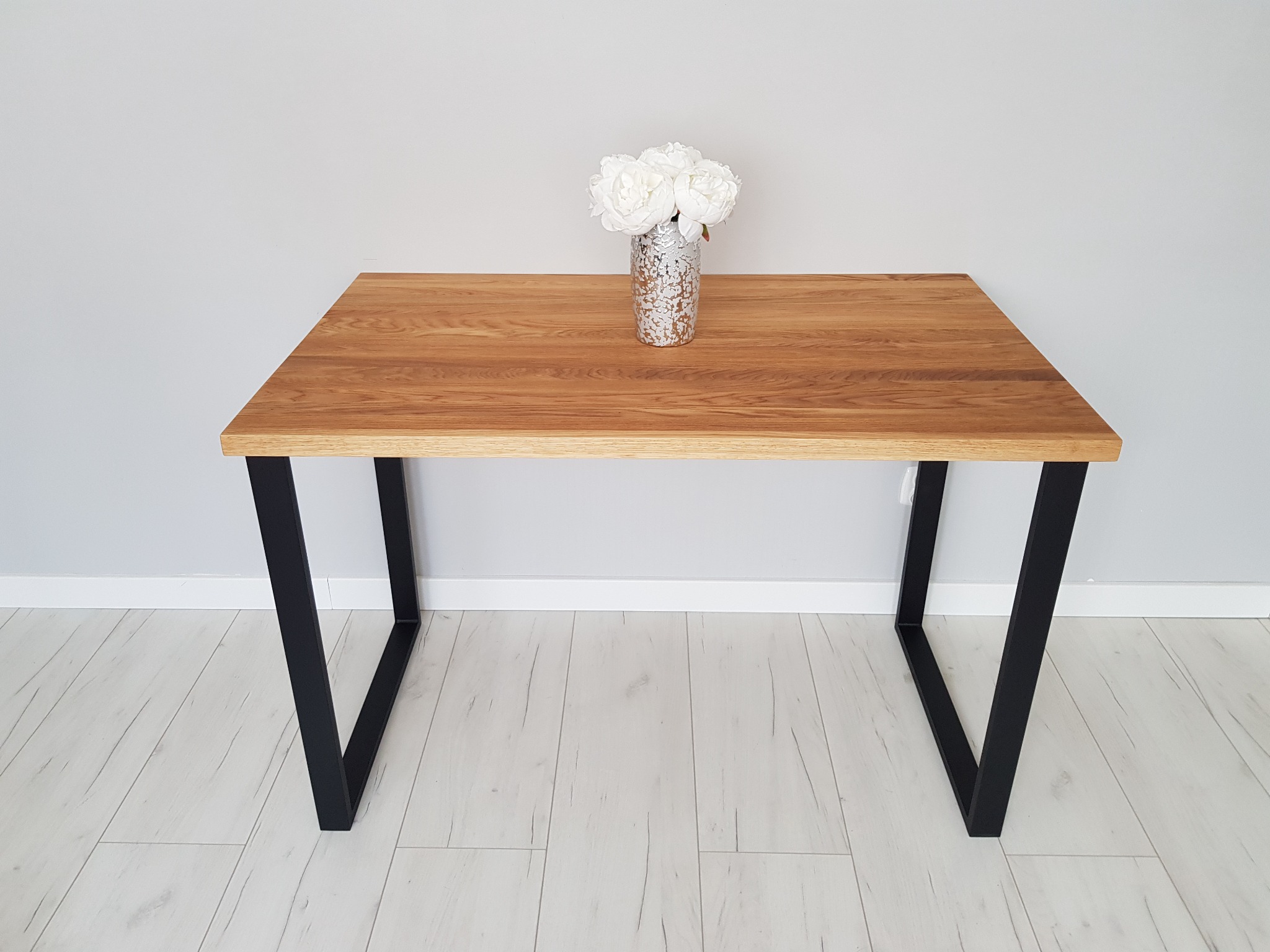 Stół z blatem z jasnego drewna i czarnymi, metalowymi nogami w kształcie litery V, na blacie wazon ze sztucznymi białymi kwiatami, na podłodze jasne panele.