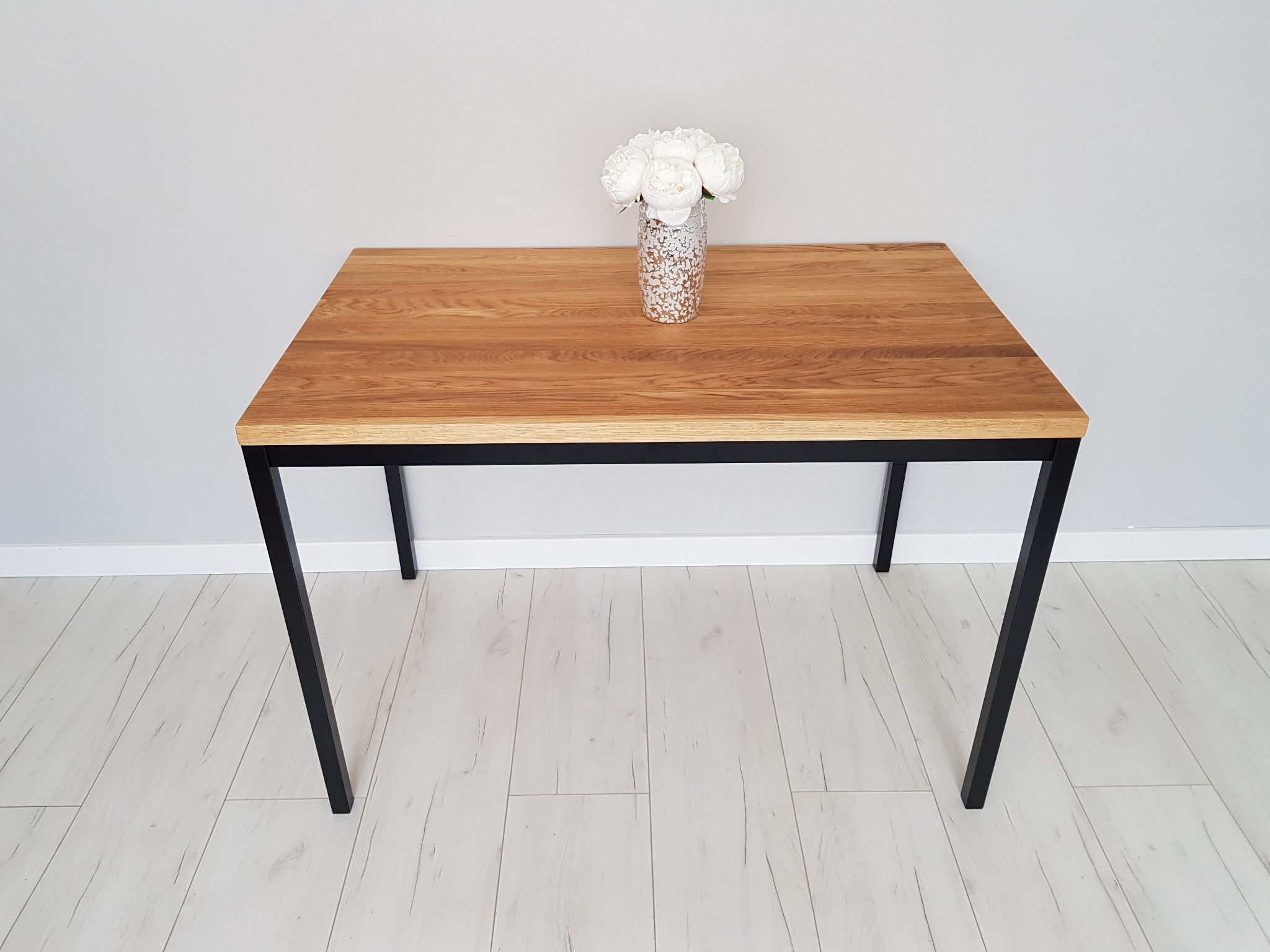 Stół z blatem z jasnego drewna i czarnymi, metalowymi nogami, na blacie wazon z białymi kwiatami, tło stanowi szara ściana i podłoga z paneli.