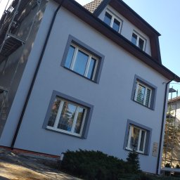 Robert Piotrowski - Dwupiętrowy dom z szarą elewacją i ciemnoszarymi obramowaniami okien, częściowo zasłonięty rusztowaniem, z widocznymi refleksami w oknach i krzewami u podstawy.